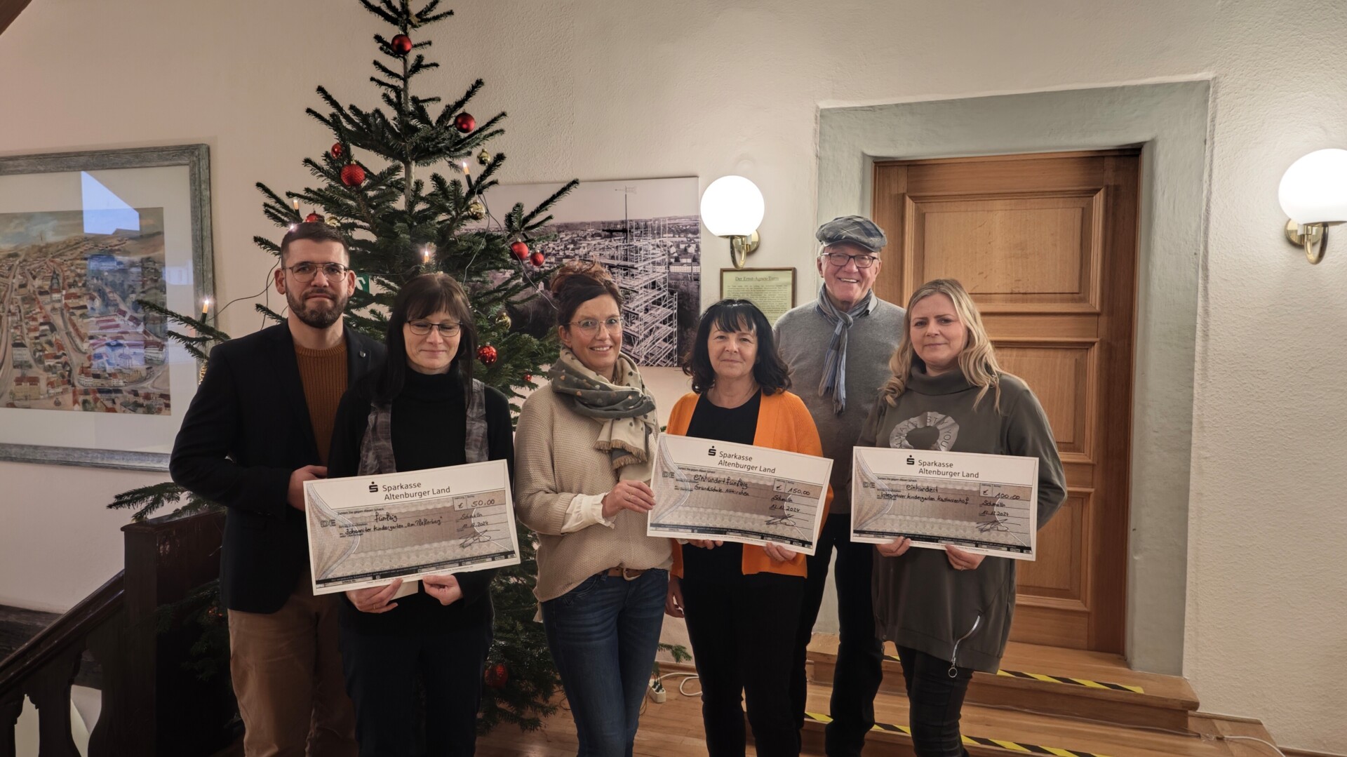 Die prämierten Einrichtungen und die Überreicher stehen mit den Spendenschecks vor einem Weihnachtsbaum, v.l.n.r.: Bürgermeister Sven Schrade, Johanniter Kita "Am Pfefferberg", Grundschule Altkirchen, Bernd Adam, Kita Finkenweg