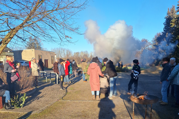 Eine Gruppe an Menschen steht beim Schneeverbrennen auf einem Platz. In einem Grill brennt Feuer, Tannenbäume werden verbrannt.